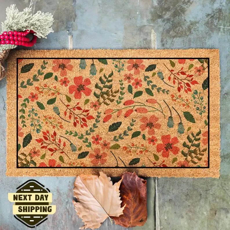 May include: A brown coir doormat with a floral pattern. The pattern features red flowers, green leaves, and small brown berries. The doormat has a black border and is displayed on a gray concrete surface. The text 'NEXT DAY SHIPPING' is visible in the lower left corner of the image.